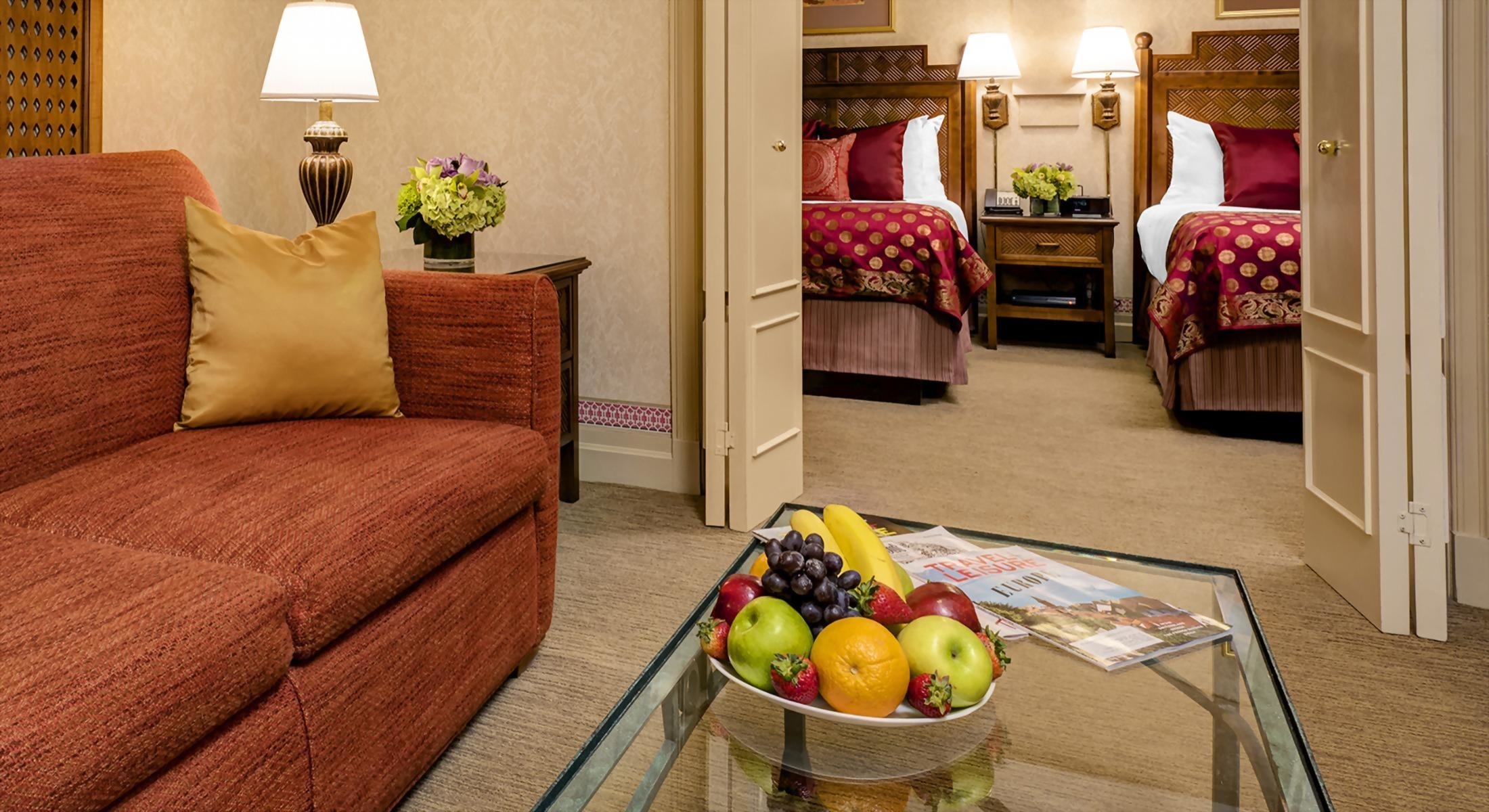 A hotel suite with a sofa, coffee table with fruit, and two beds visible through a doorway.